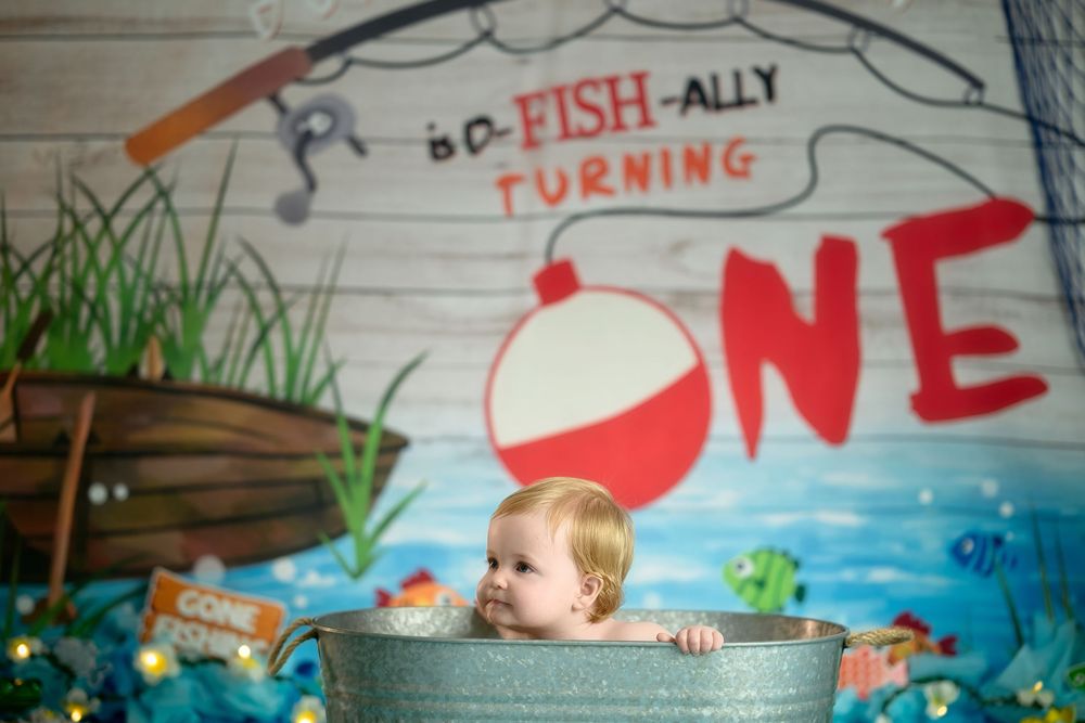 A fishing-themed first birthday party setup with a child sitting in a galvanized tub against a painted ocean backdrop.
