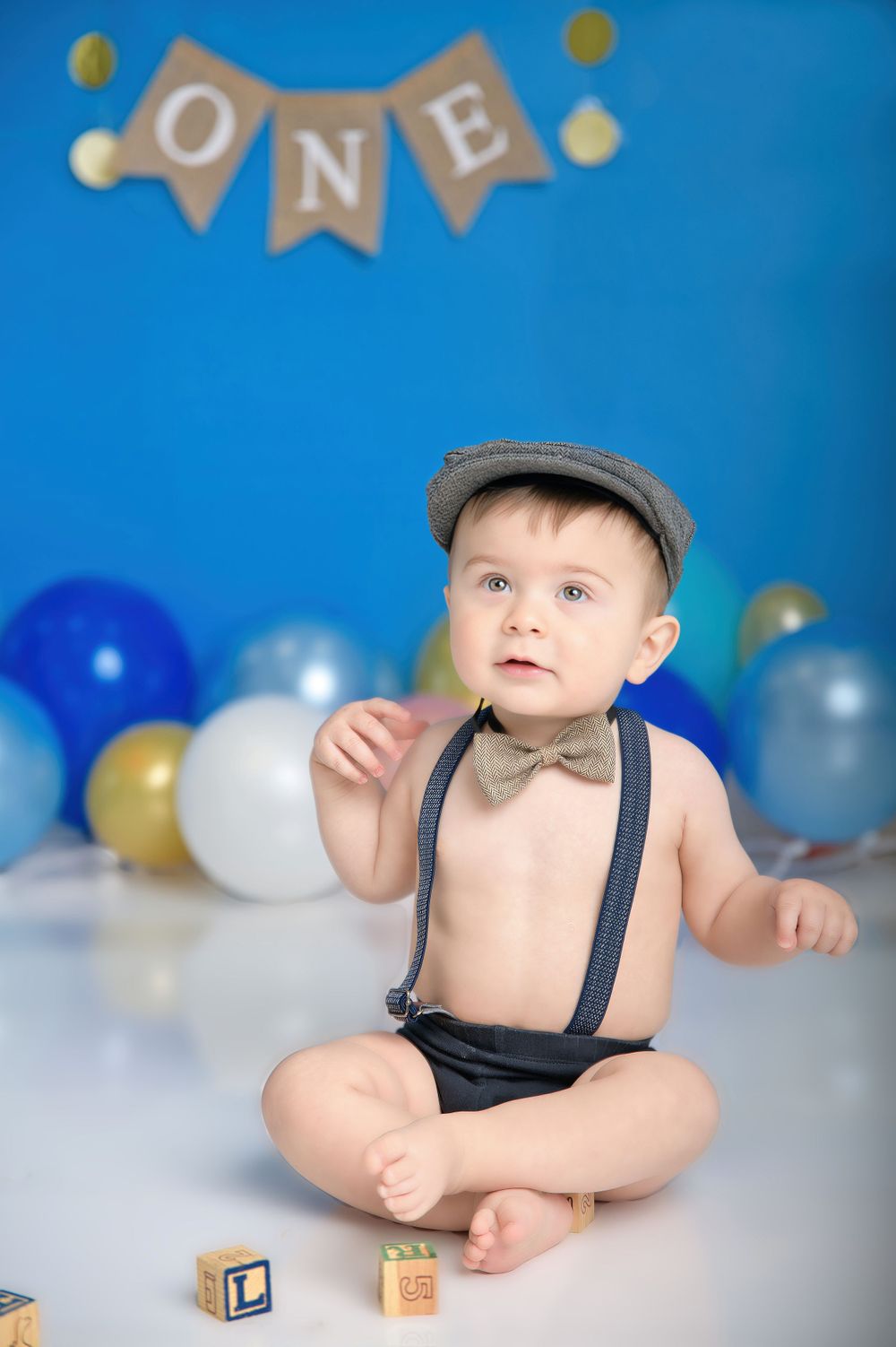 First birthday photo shoot setup with balloons, wooden blocks, and vintage-style suspenders and cap against blue backdrop.