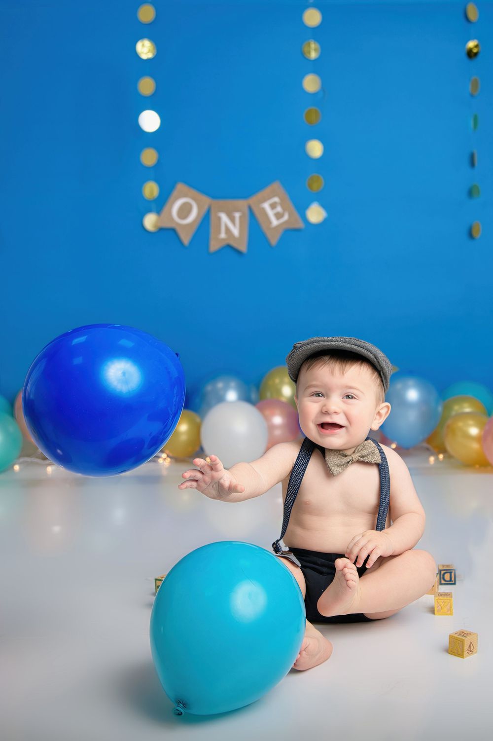 First birthday photoshoot setup with blue and gold balloons, suspenders, and 'ONE' banner against blue backdrop.