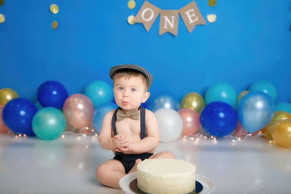 First birthday celebration setup with colorful balloons, ONE banner, and smash cake against blue backdrop.