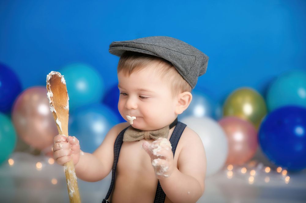 A toddler in a newsboy cap and bow tie holds an ice cream cone against a festive backdrop of blue and silver balloons.