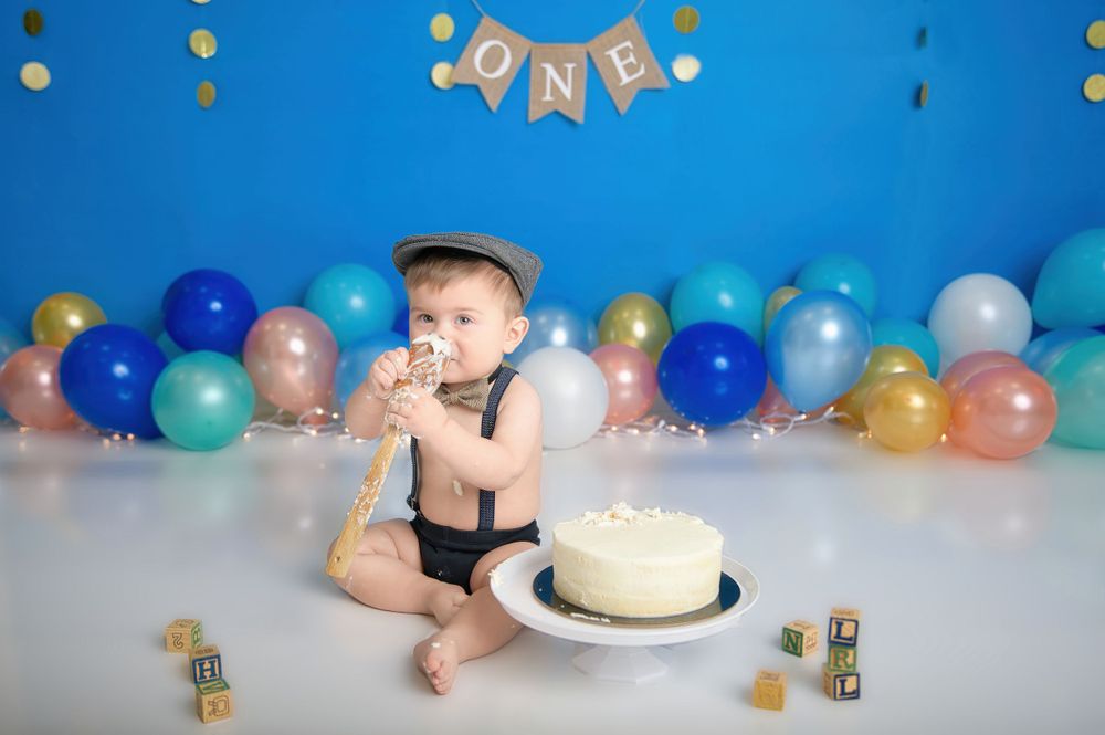 A cake smash first birthday photo setup with blue and gold balloons against a blue backdrop and a white frosted cake.