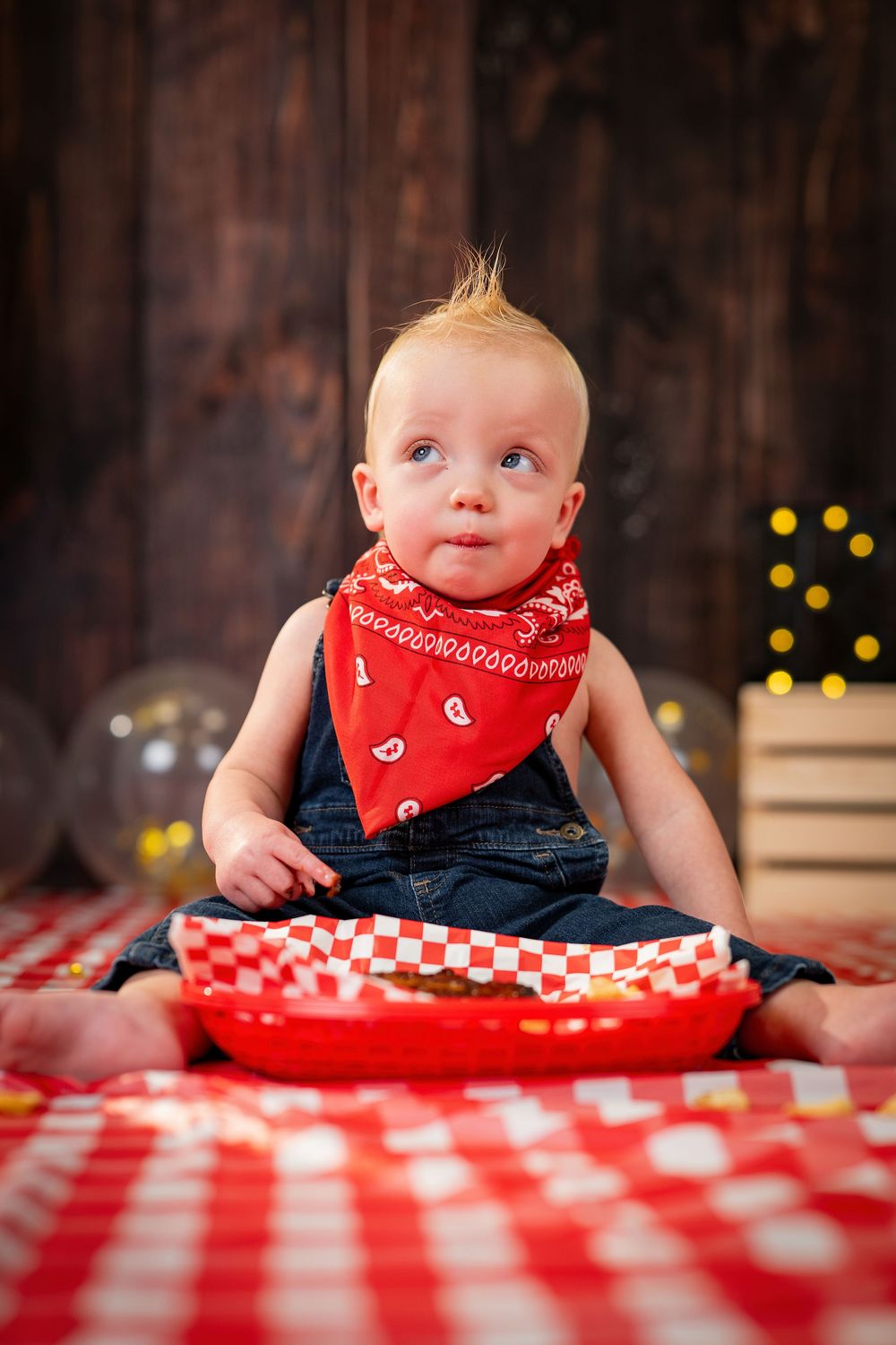 Baby in red bandana sits at table with red and white checkered tablecloth and glowing lights in background.