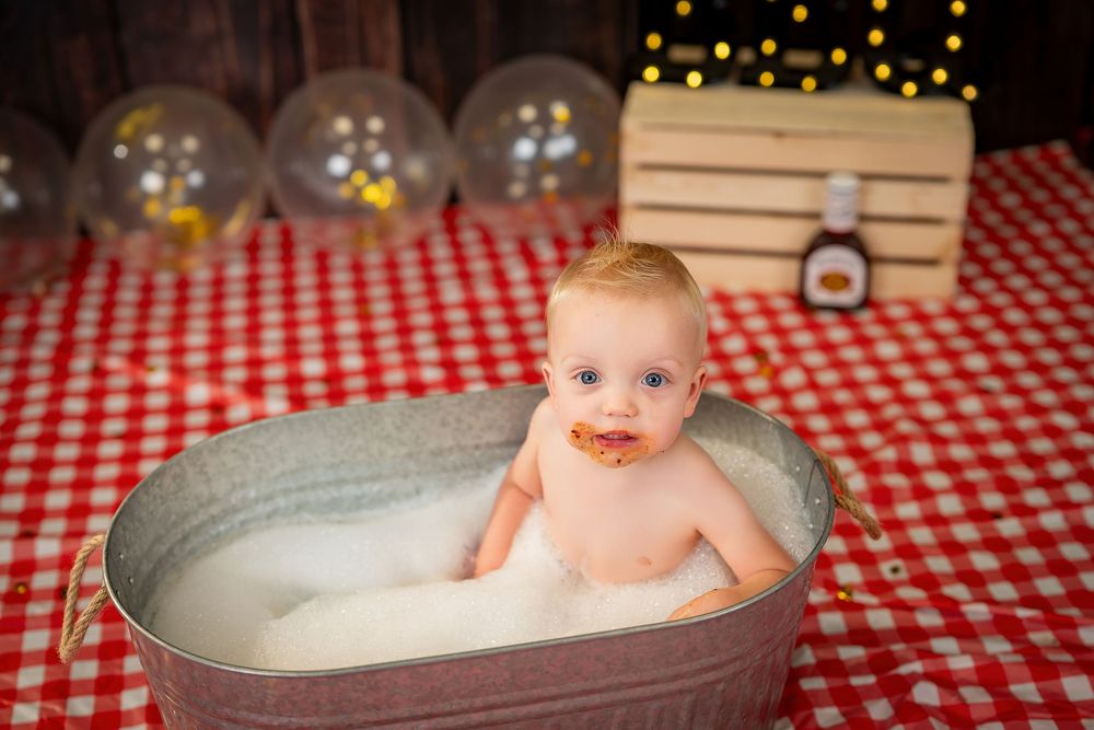 Baby wearing red bandana bib sits in front of red and white checkered tablecloth with twinkling lights behind.