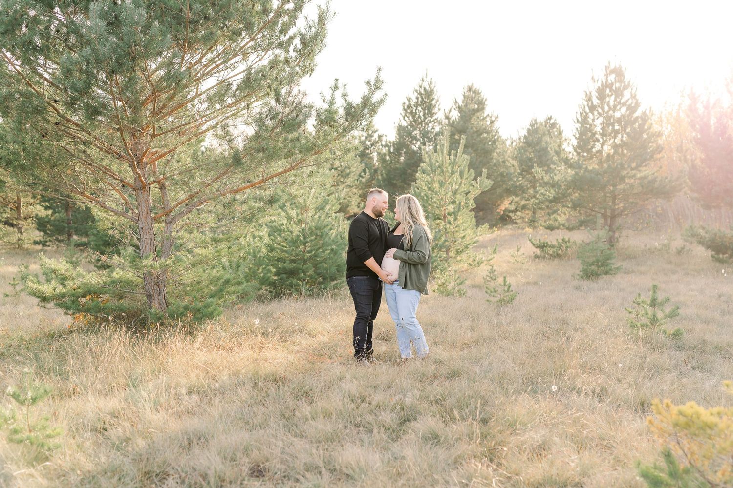 A couple stands together in a sunlit field surrounded by evergreen trees during golden hour.