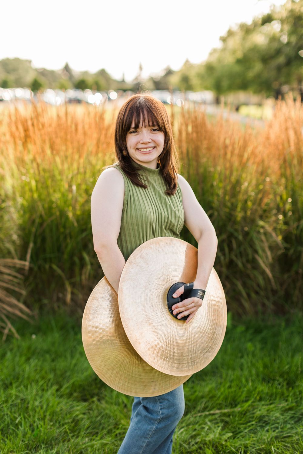 A person in a green sleeveless top holds cymbals while standing in front of tall grasses at sunset. Twin cities senior photography.