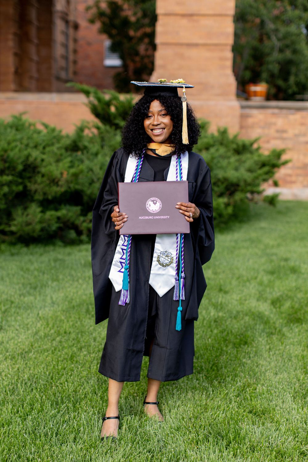 A graduate in black cap and gown stands on grass holding a diploma while wearing graduation stoles. Twin cities senior photography.