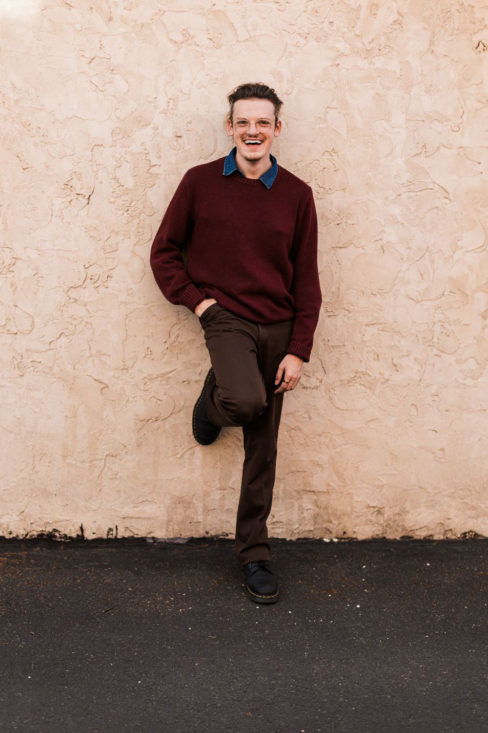 Someone in a burgundy sweater and dark pants stands casually against a textured beige wall. Twin cities senior photography.