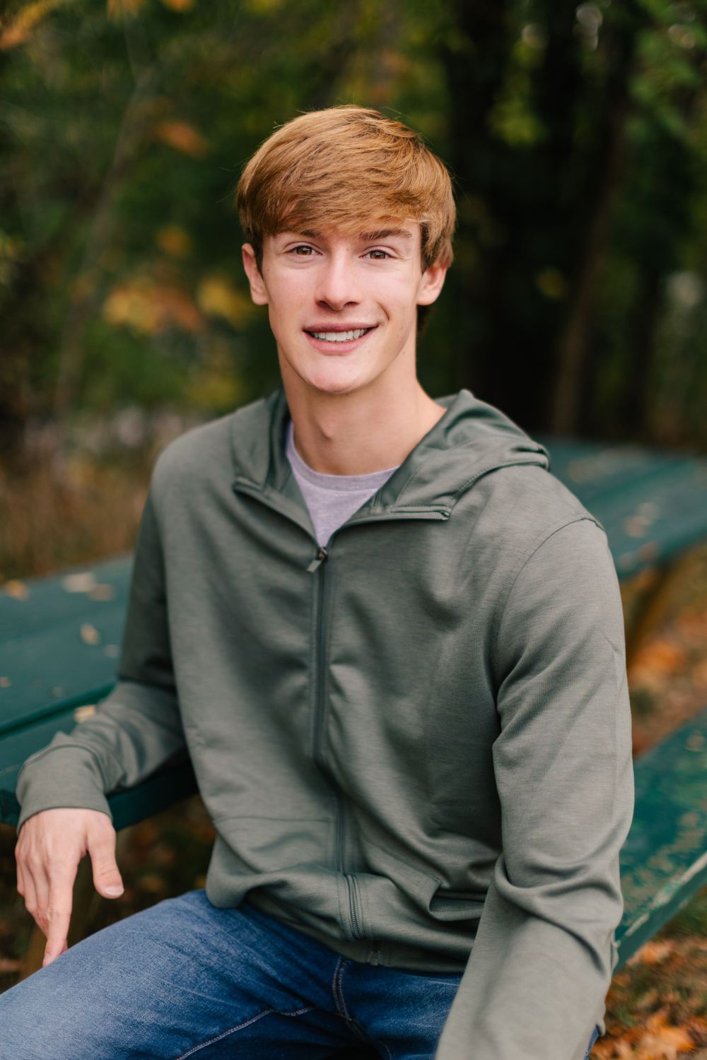 Young person with auburn hair wearing a gray hoodie and jeans sits smiling outdoors on a bench in autumn. Twin cities senior photography.