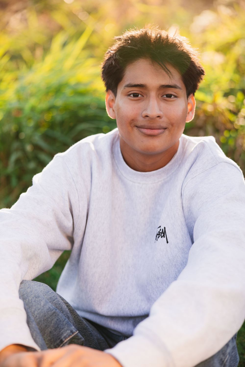 Person in a white sweatshirt sitting outdoors with sunlit foliage in the background during golden hour. Twin Cities senior photography.