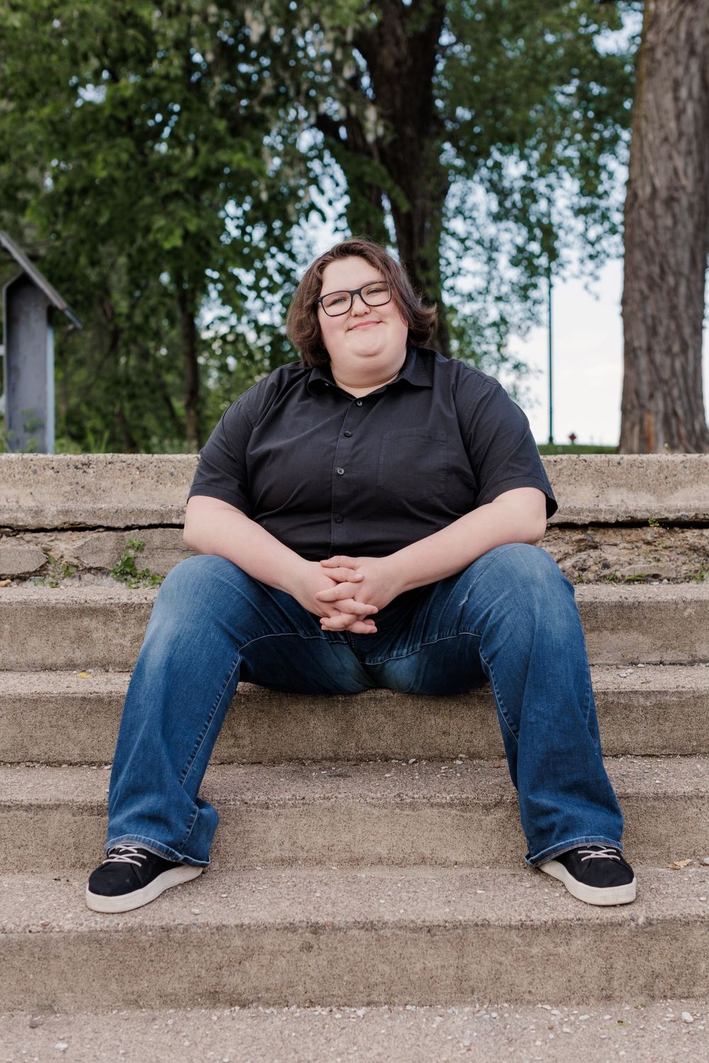 Someone sits casually on concrete steps wearing a black polo shirt, jeans and glasses on a sunny day. Twin cities senior photography.