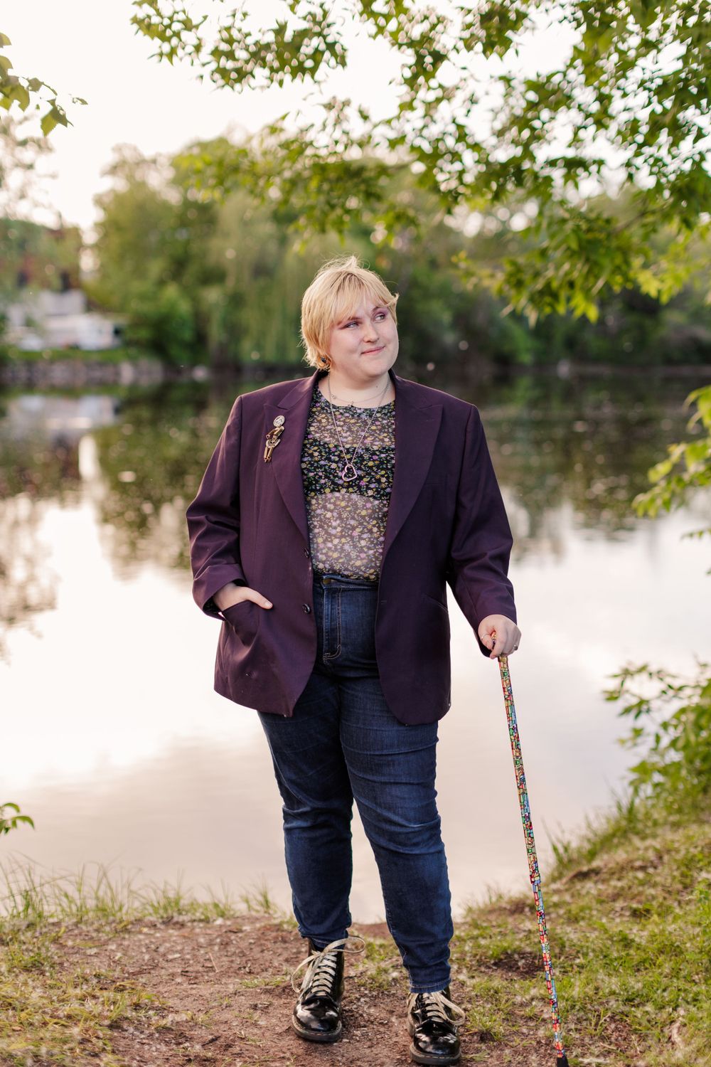 A person wearing a purple blazer and navy pants leans on a walking cane by a tranquil lake at dusk. Twin cities senior photography.