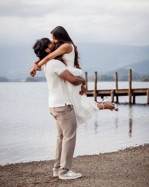 Couple embracing and kissing by a lakeside dock, dressed in white and beige outfits, with mountains in the background.