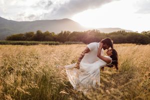 Couple sharing a romantic dip kiss in a golden wheat field with mountains and sunset in the background.