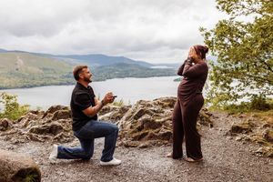 Man proposes on one knee to woman at scenic loch overlook with mountains and water in background.