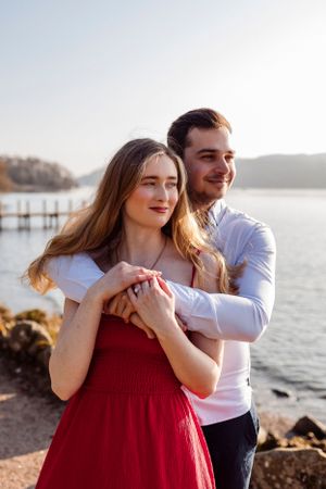 A romantic couple embraces by the water at sunset, with a dock and rocky shore visible in the background.
