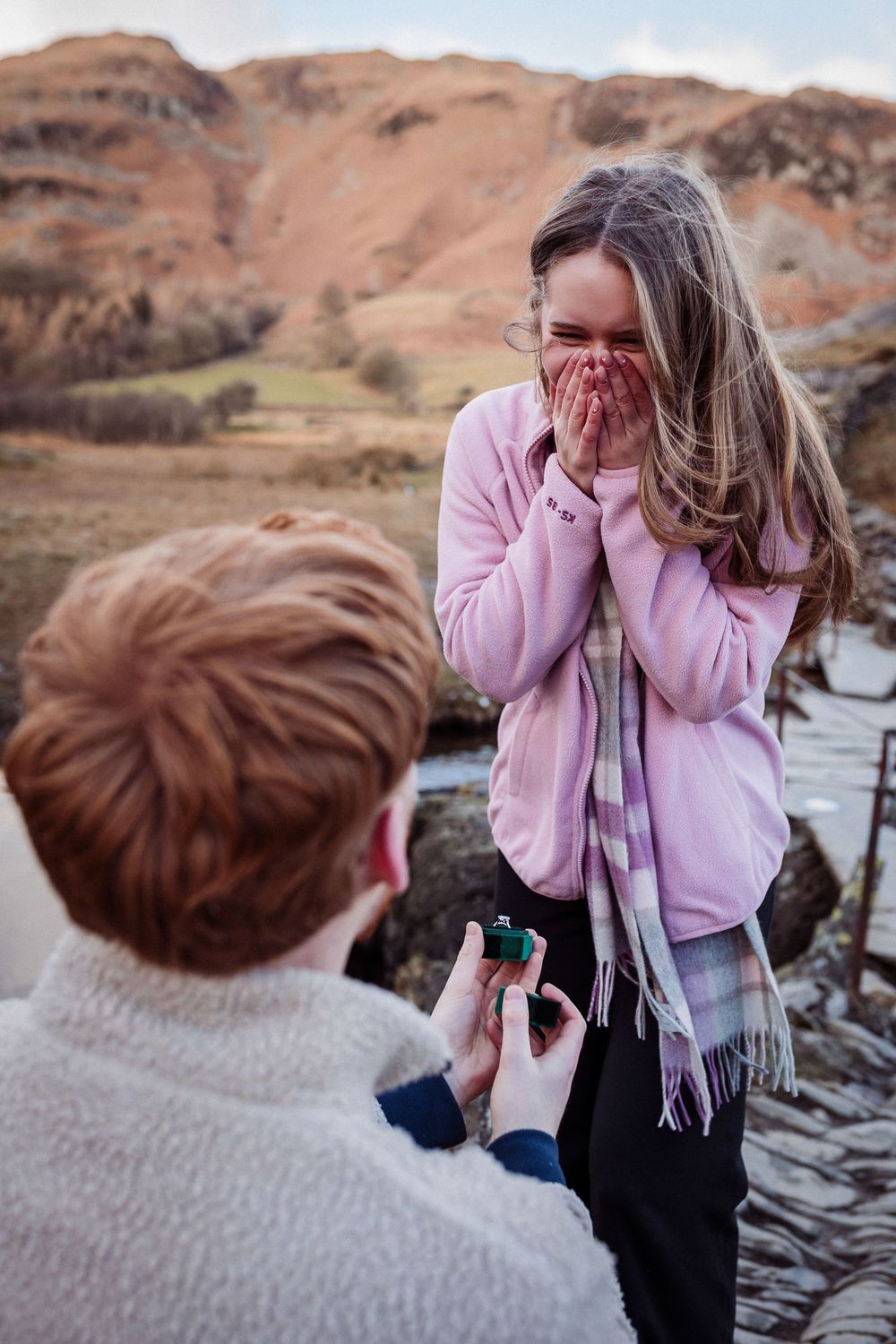 A surprise proposal moment captured at a scenic rocky location with someone in a pink sweater reacting with joy.