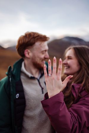 A couple sharing a romantic moment outdoors while showing off an engagement ring on an outstretched hand.