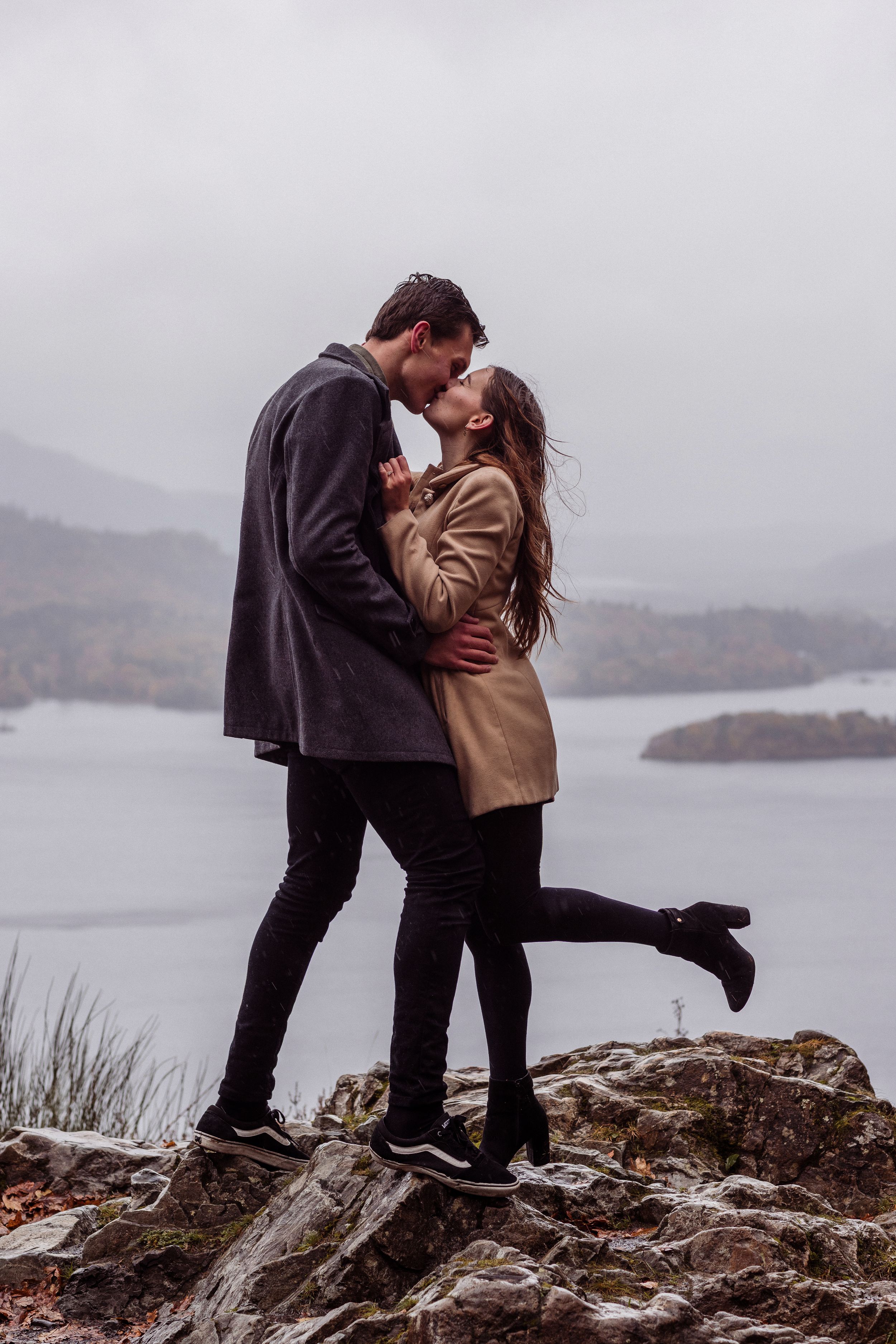 A couple shares a romantic kiss while standing on rocky cliffs overlooking a misty lake landscape.