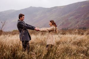 A couple holds hands while standing in a field of tall grass with misty mountains in the background.