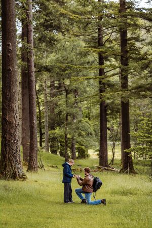 Man proposing on one knee to partner in a lush green forest clearing surrounded by tall pine trees.