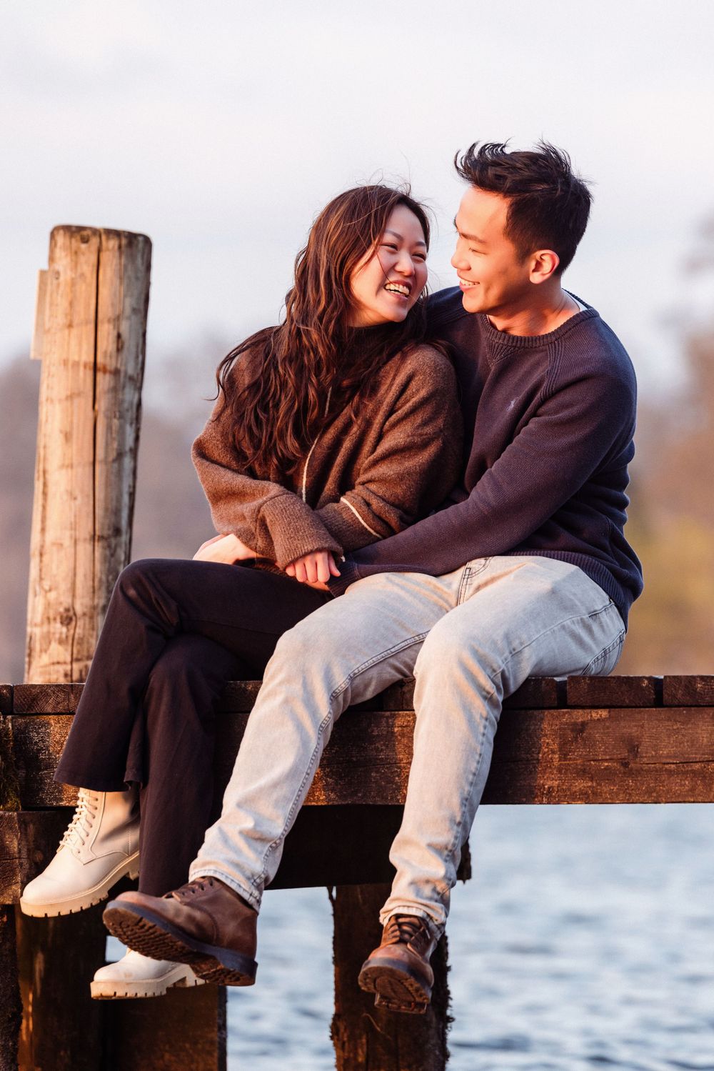 A couple shares a joyful moment sitting together on a wooden dock by the water during sunset.