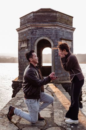 A couple shares a romantic marriage proposal moment at sunset near a stone lighthouse structure by the water.