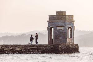Two silhouettes stand together at a historic stone pavilion overlooking a misty waterfront at sunrise at Storrs Hall, Temple of Heroes.
