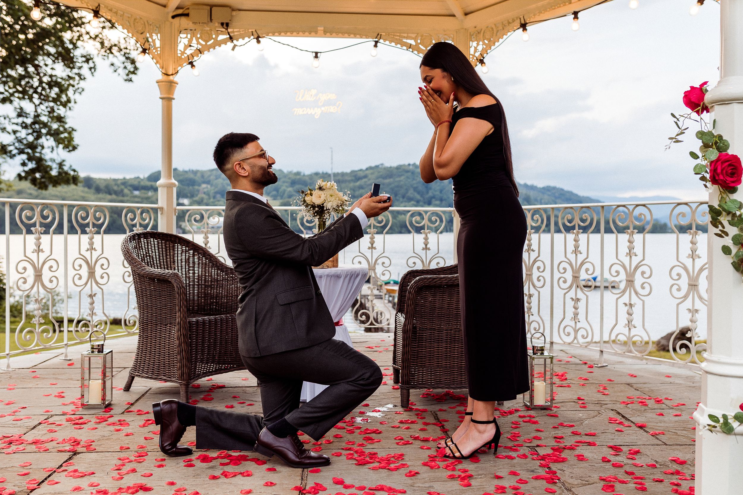 Man proposes to woman on one knee under gazebo with rose petals scattered on ground during romantic engagement.