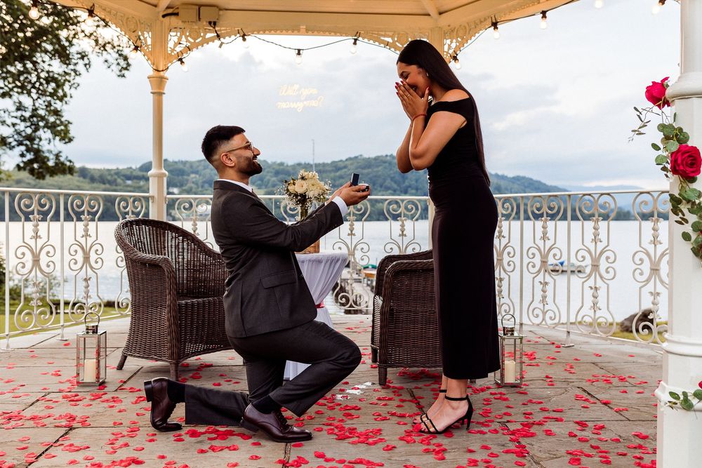 Man proposes to woman on one knee under gazebo with rose petals scattered on ground during romantic engagement.