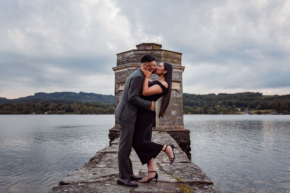 Couple sharing a romantic kiss on a stone pier by a lake with a historic tower and cloudy skies backdrop.