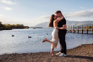 Couple embracing and kissing by a lakeside on a pebble beach with mountains and a pier in the background.