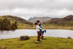 A couple embraces in a scenic mountain landscape with a serene lake and rolling hills in the English countryside.