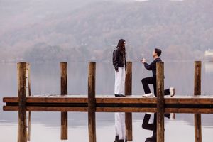 Two people walk along a wooden pier on a foggy morning with mountains visible in the distance across the lake.
