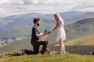 A couple shares a romantic moment on a scenic mountaintop overlooking rolling hills and valleys in the distance.