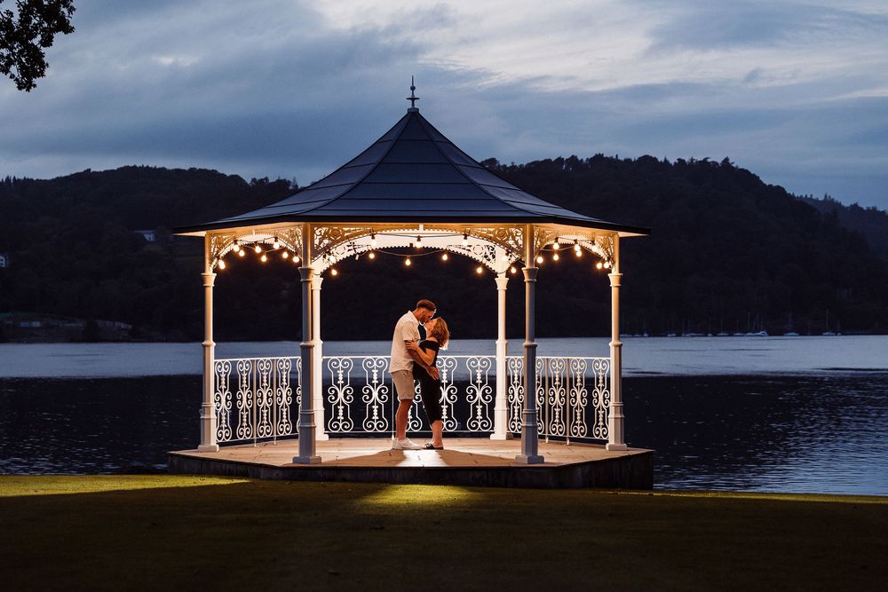 A romantic gazebo with string lights illuminated at dusk by a lake with mountains in the background.