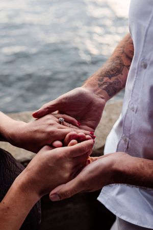 Close-up of hands during marriage proposal by the ocean with diamond ring and blurred sparkling water background.