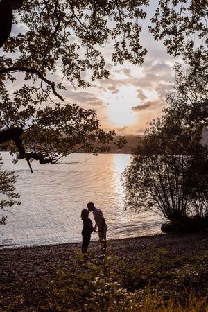 Two silhouettes stand together at the water's edge during a serene sunset, framed by overhanging tree branches.