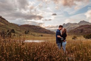 Two people share an intimate moment at sunset in the mountains surrounded by rustic field grass and dramatic clouds.