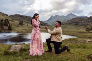 Man proposing to woman in floral dress by a lake with mountains in the background.