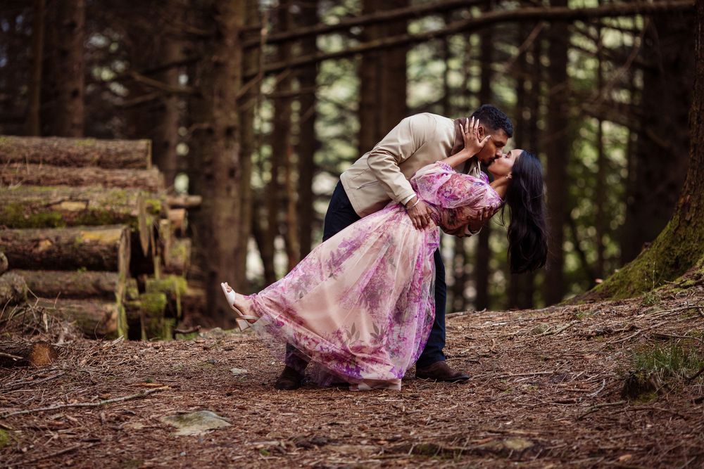 Couple sharing a romantic dip kiss in a misty forest, woman in flowing pink gown, surrounded by tall pine trees.