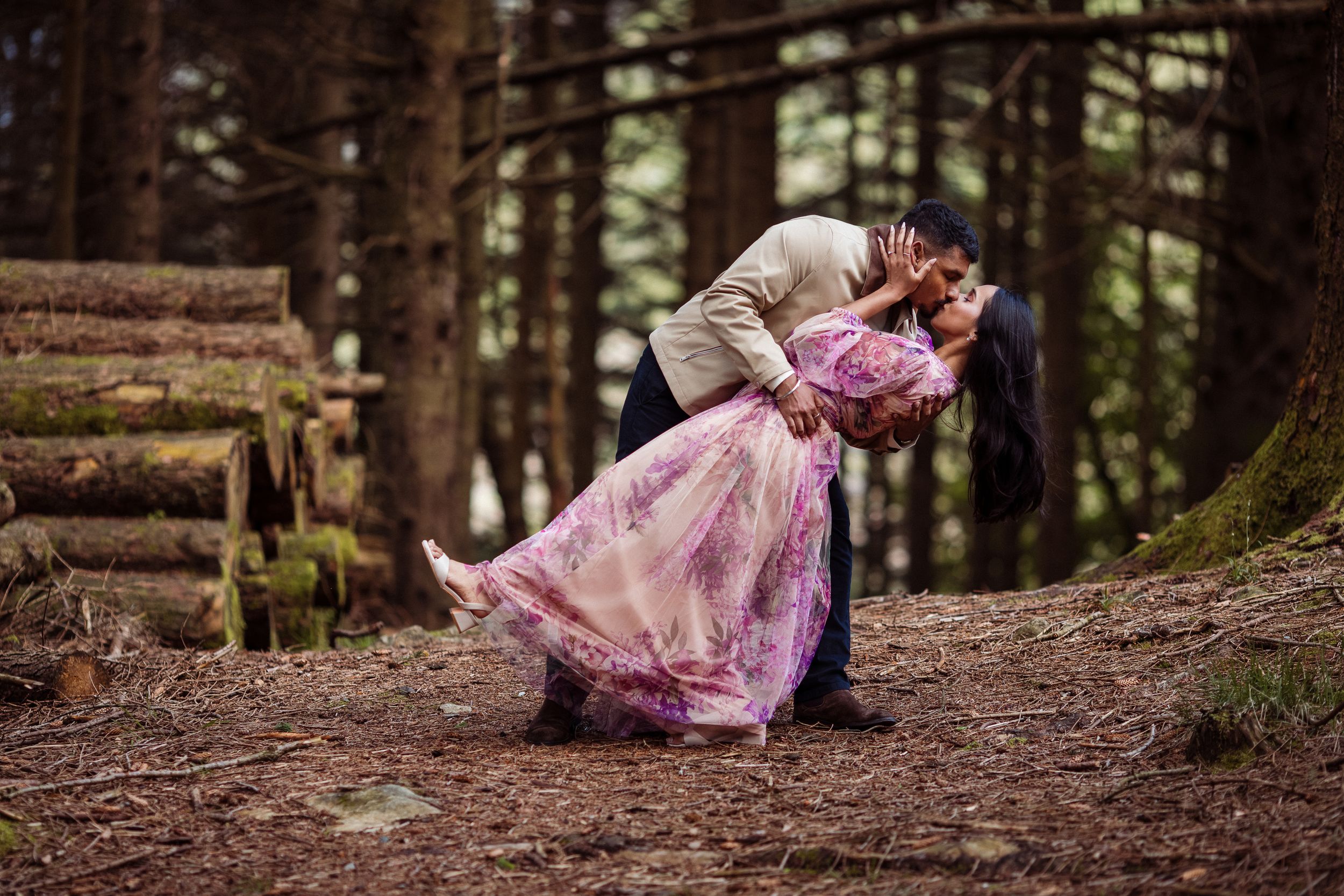 Couple sharing a romantic dip kiss in a misty forest, woman in flowing pink gown, surrounded by tall pine trees.