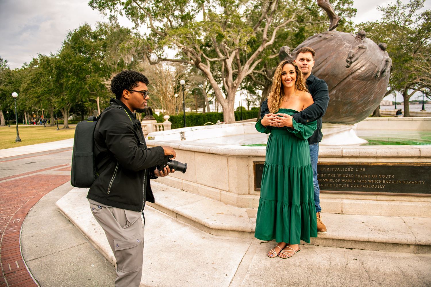 A photographer captures a couple posing near a stone monument in a park on a sunny day.