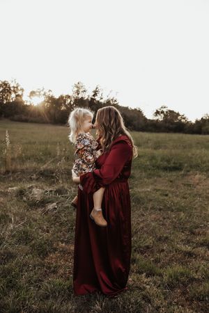 Mother in burgundy dress holding toddler daughter nose-to-nose in golden sunset field.
