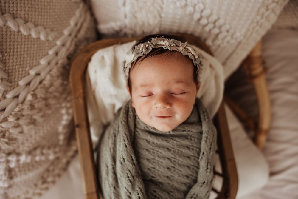 Peaceful sleeping newborn swaddled in sage green knit wrap wearing delicate floral headband in bowl.