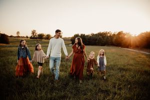 Family of six holding hands walking through green field together at golden hour sunset.