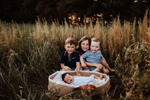 A mother sits in a golden field with three young children, including a newborn in a basket, surrounded by tall grass.