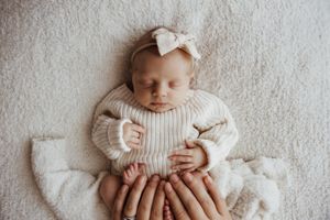 Peaceful newborn baby in a cream knit outfit with a bow headband, cradled by family hands on a soft blanket.