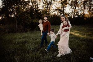 A joyful family of five plays in a lush green field at sunset, with parents holding young children.
