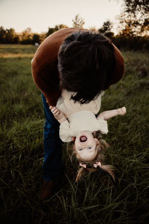 A parent playfully holds a laughing toddler upside down in a green field during golden hour.